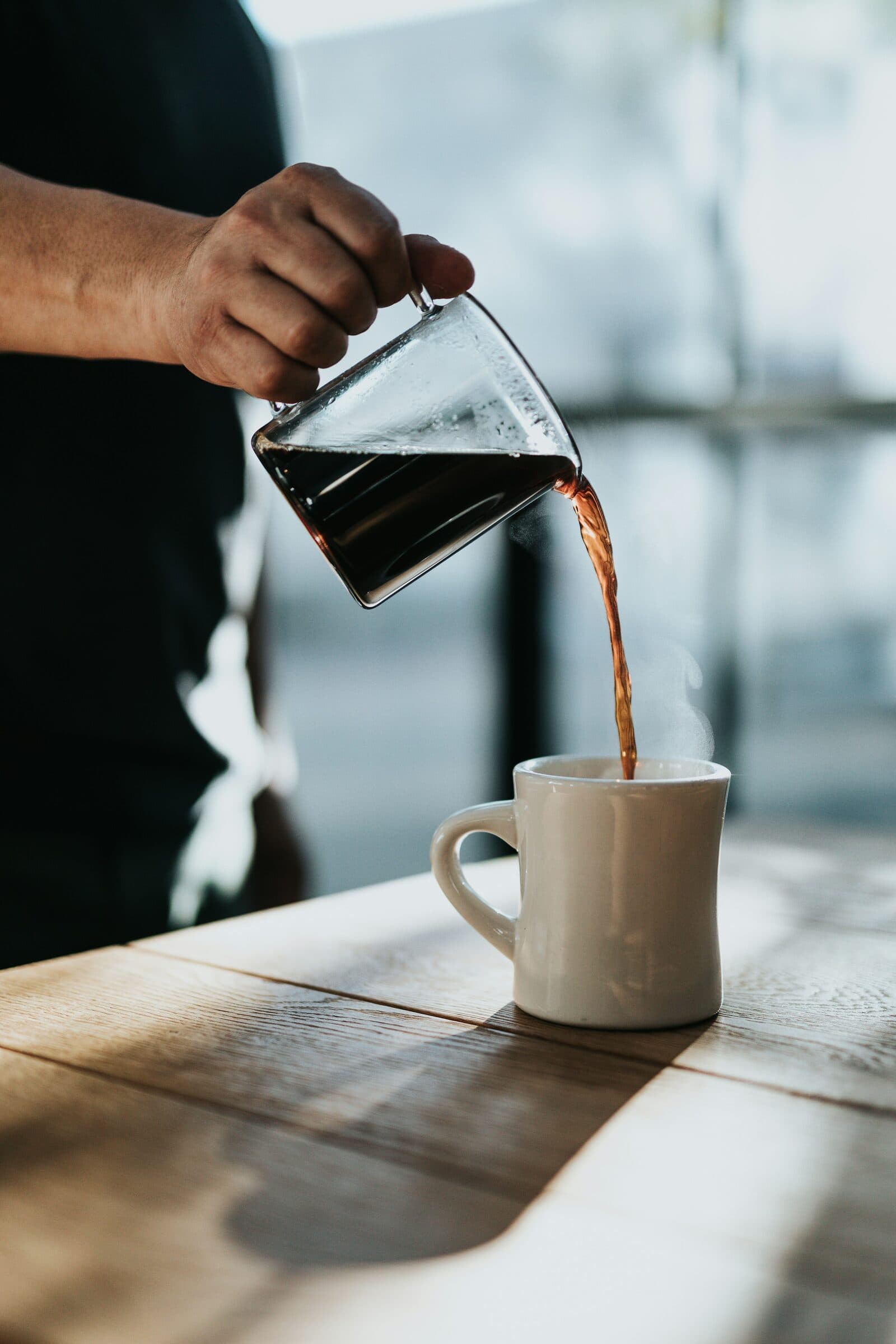 Hot coffee poured from a glass carafe into a white mug by a window.
