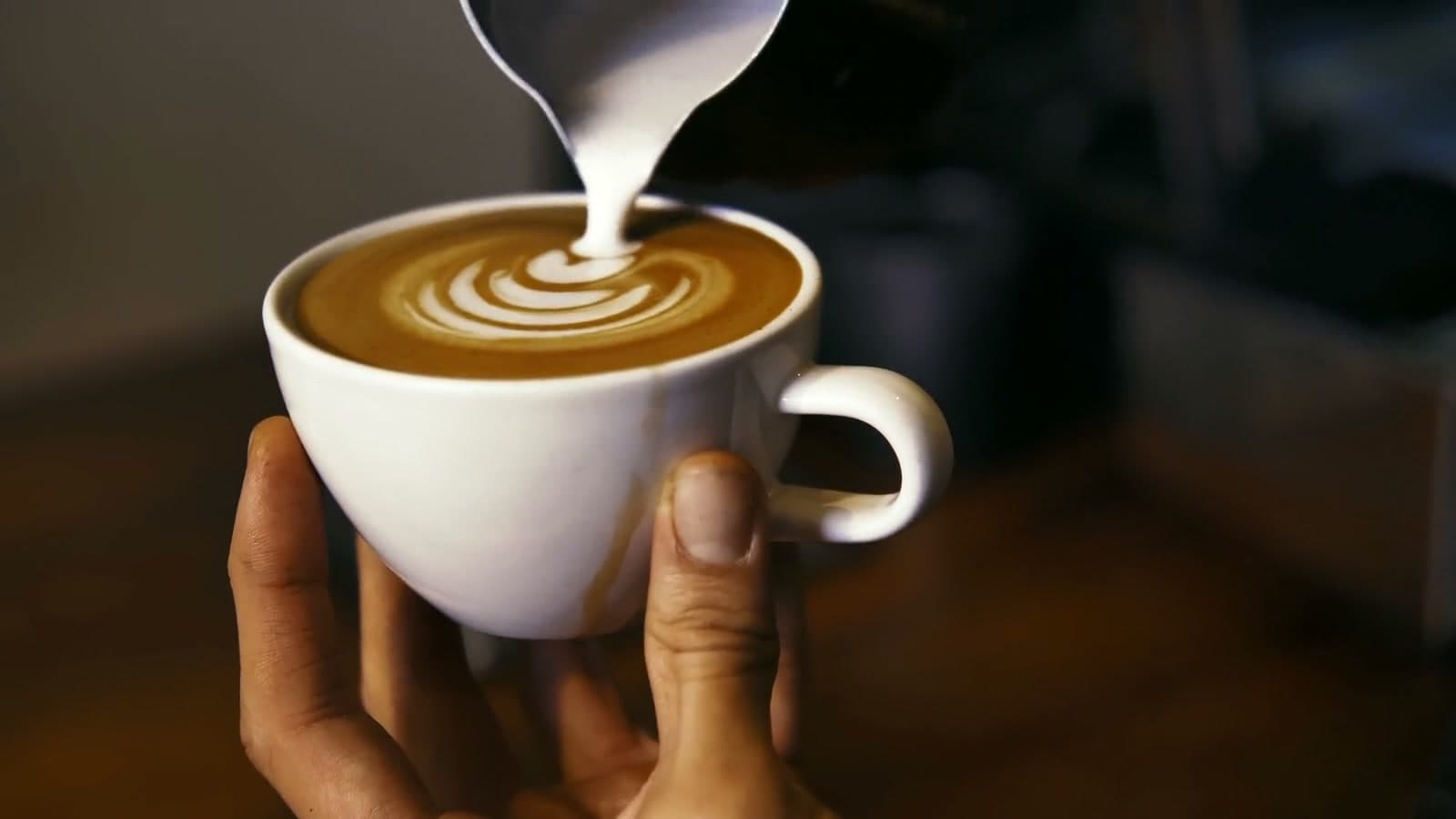 Hands hold a finished latte while a little more milk is poured in.