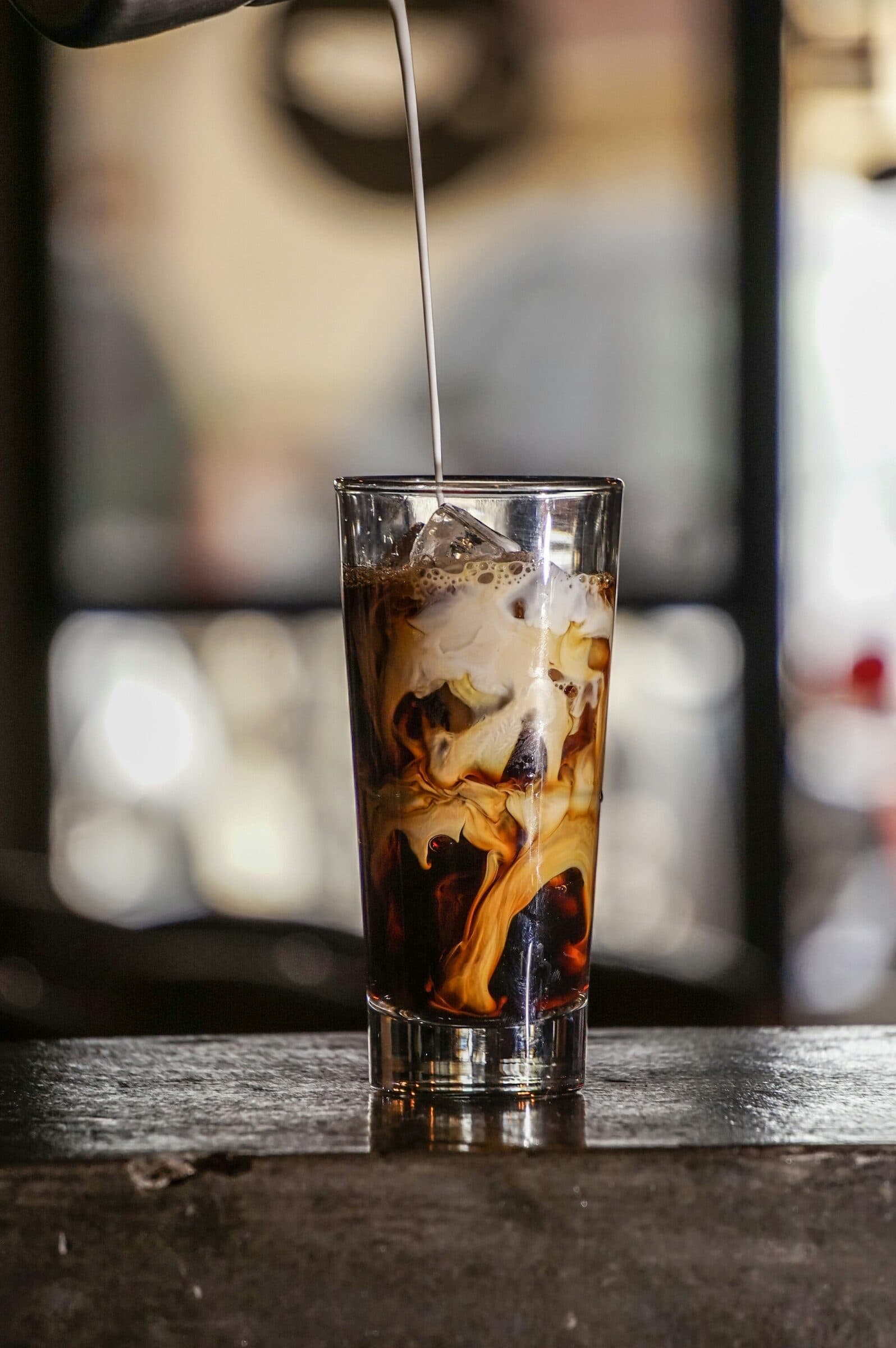 Milk pouring into a tall glass of iced coffee, swirling through the ice.