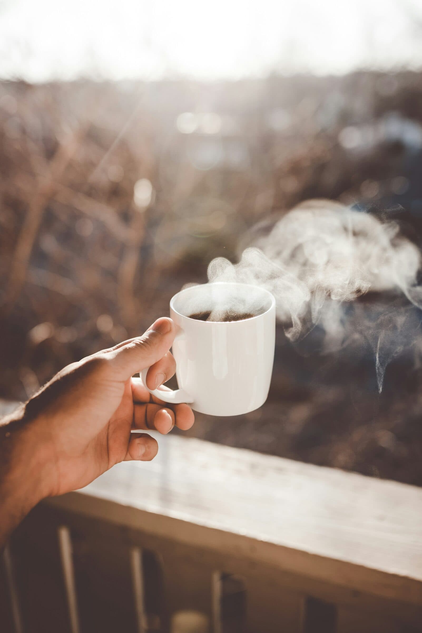 A hand holds a steaming white mug outdoors on a cold morning.