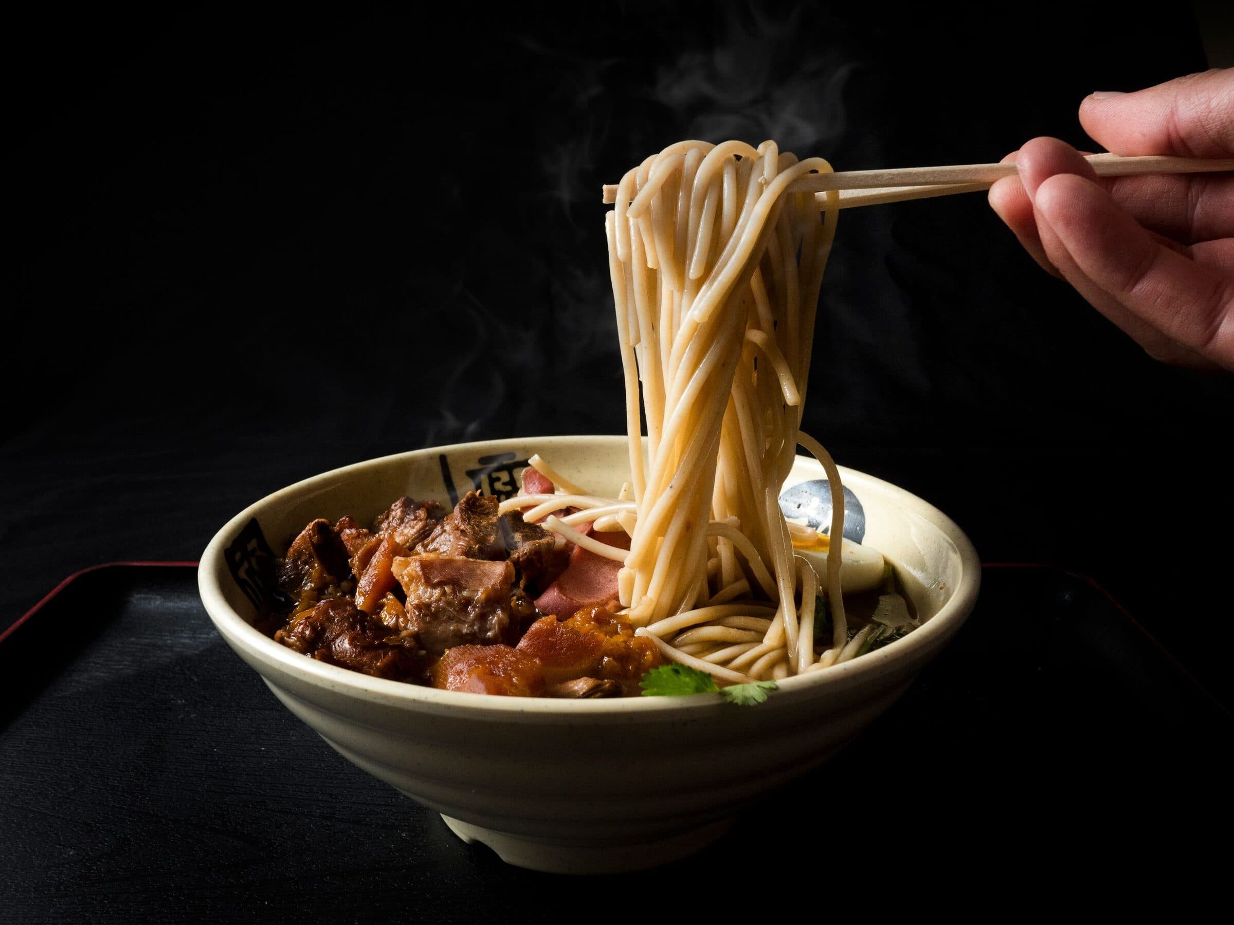 Noodles lifted from a steaming bowl of ramen with chopsticks.