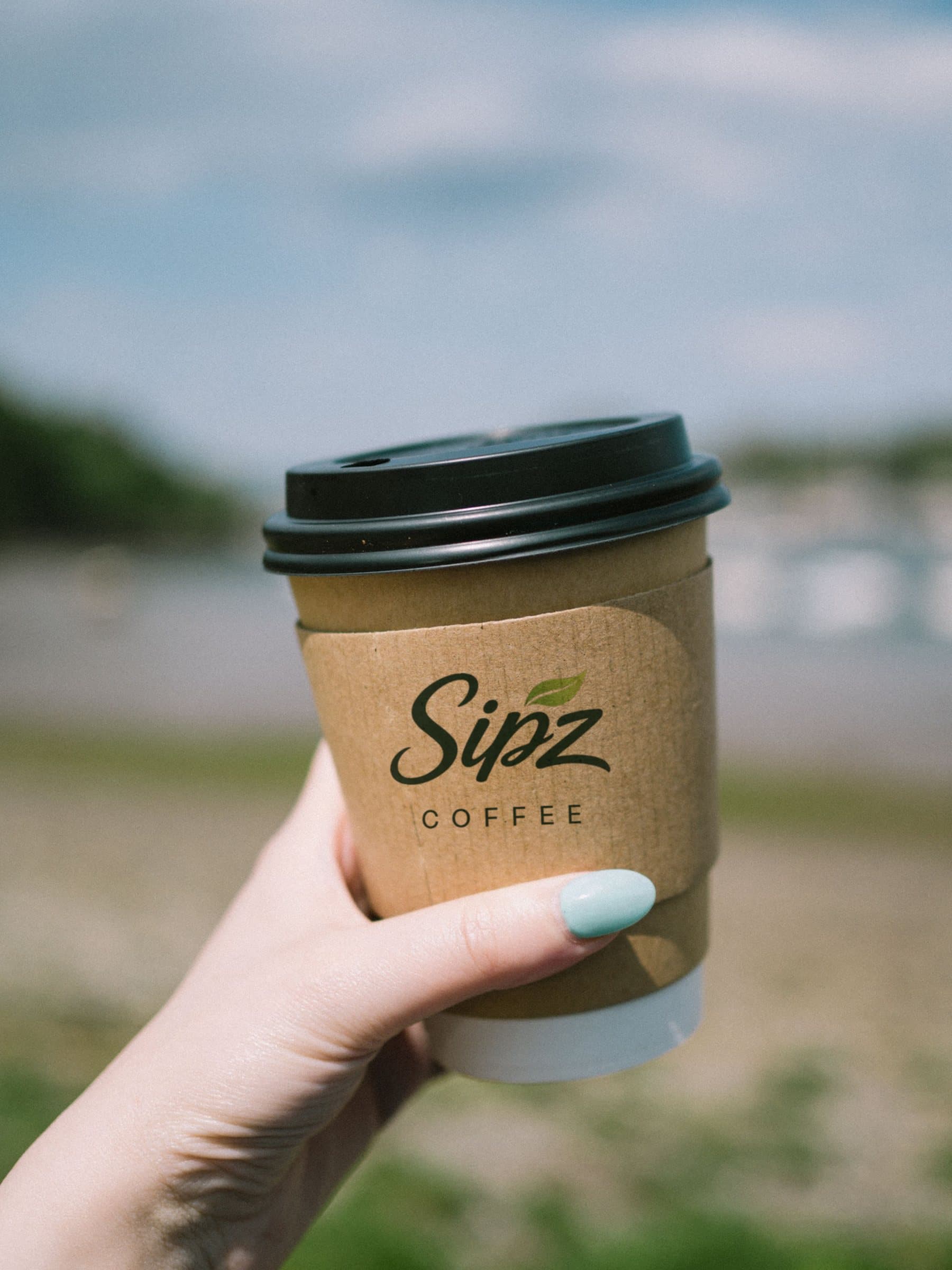 A hand holds a SIPZ coffee in a kraft sleeve outdoors on a sunny day.