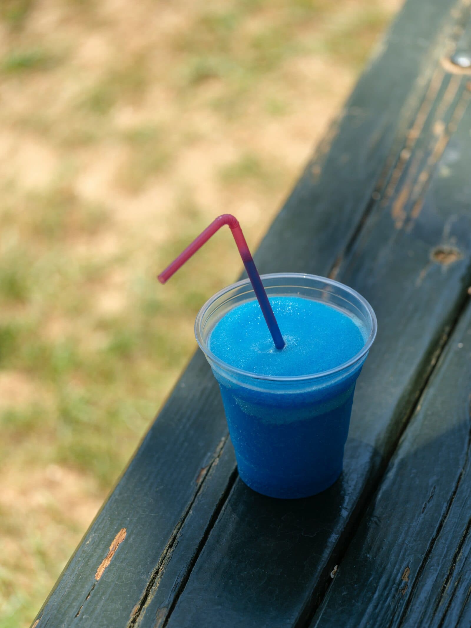 A blue raspberry slush with a bendy straw, on a wooden picnic bench.