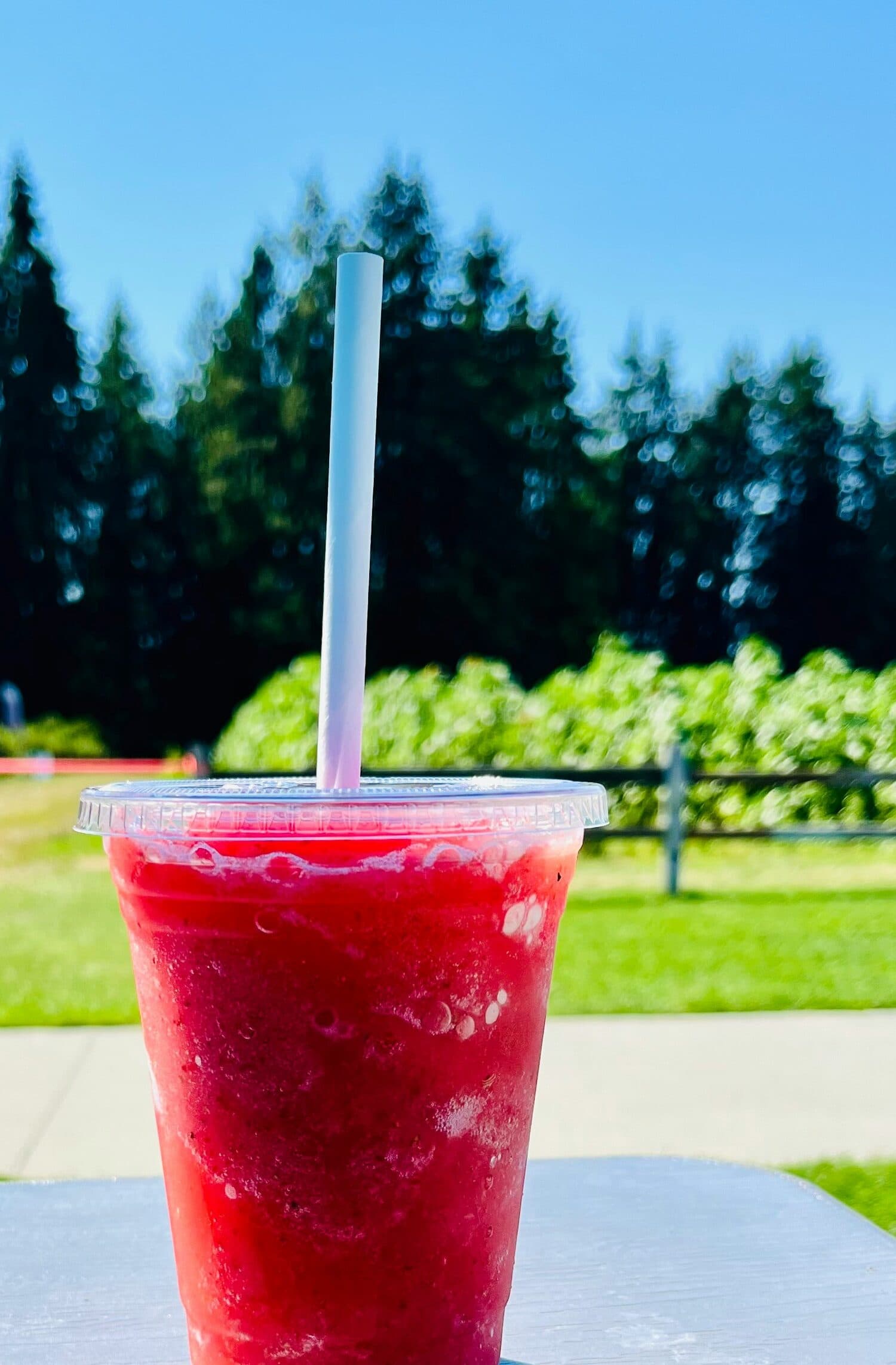 A red slush in a clear to-go cup with a straw, on a picnic table on a sunny summer day.